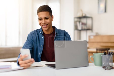 Foto de Un joven macho sonríe contento mientras mira su teléfono inteligente, con un portátil abierto en el escritorio frente a él en un interior acogedor - Imagen libre de derechos