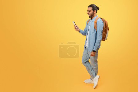 Foto de Hombre indio casual con mochila caminando y mirando el teléfono inteligente sobre un fondo amarillo - Imagen libre de derechos