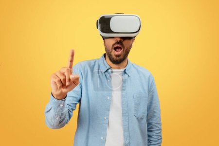 Indian man in casual attire is engaged with a virtual reality headset, pointing upwards, on a mustard background