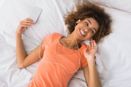 Téléchargez les photos : Une jeune femme afro-américaine s'allonge sur son lit tenant un smartphone, avec des écouteurs connectés, suggérant détente ou temps libre à la maison - en image libre de droit