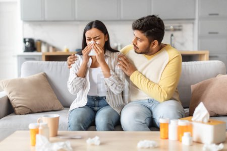 Foto de El hombre indio consuela a una mujer enferma que estornuda en un pañuelo, rodeada de remedios fríos en casa.. - Imagen libre de derechos
