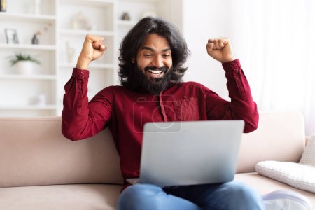 Ein überschwänglicher indischer Mann sitzt auf einer Couch, die Arme im Sieg erhoben, und blickt auf einen Laptop-Bildschirm, in einer häuslichen Umgebung