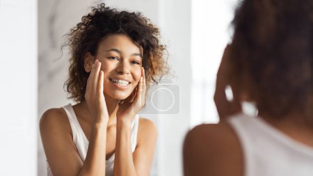 Téléchargez les photos : Joyeux afro fille touchant sur son visage, debout dans la salle de bain, réfléchissant dans le miroir. Concept beauté et soins - en image libre de droit