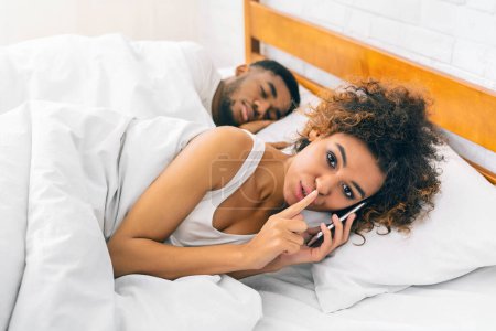 Photo for African American young woman lies in bed talking on her phone while a man sleeps beside her, hinting at modern relationship dynamics - Royalty Free Image
