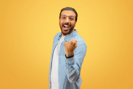 Téléchargez les photos : Exuberant homme indien barbu avec des lunettes soulève son poing dans l'excitation sur un fond jaune - en image libre de droit