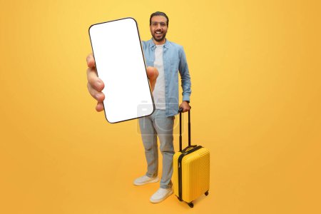 Téléchargez les photos : Un homme indien joyeux en tenue décontractée tenant un smartphone avec un écran vierge et une valise jaune sur fond jaune - en image libre de droit