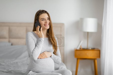 Téléchargez les photos : Jeune femme enceinte parlant sur un téléphone portable avec une expression joyeuse et la main sur le ventre - en image libre de droit
