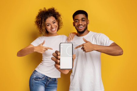 Téléchargez les photos : Un jeune couple afro-américain joyeux pointe vers un écran de smartphone vierge, parfait pour afficher des applications ou des publicités - en image libre de droit