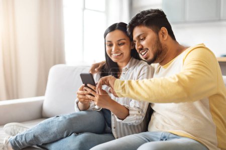 Photo for Indian man and woman are seated on a couch, both looking intently at a cell phone screen, couple relaxing at home - Royalty Free Image