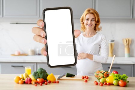 Téléchargez les photos : Femme blonde enceinte tenant un écran de smartphone vide dans une cuisine avec des produits frais - en image libre de droit