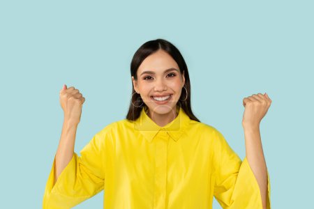 Photo for Gestures and emotions. Emotional happy beautiful young latin woman in casual outfit clenching fists and grimacing, celebrating success on blue studio background, got good news, copy space - Royalty Free Image