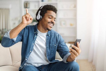 Homme noir joyeux assis sur un canapé, absorbé par l'écoute de musique sur son smartphone.