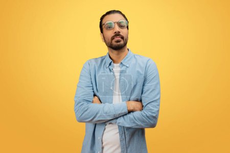 A self-assured indian man with a beard and glasses stands with crossed arms in a denim shirt, against a yellow backdrop