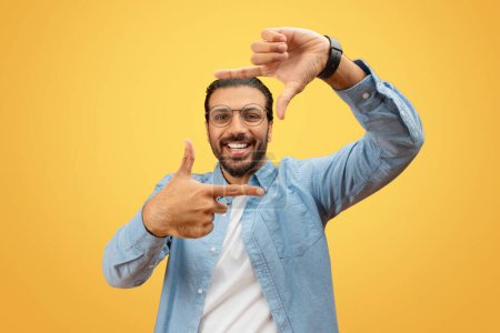 Fröhlicher indischer Mann mit Brille umrahmt sein Gesicht mit den Händen vor einem schlichten gelben Hintergrund