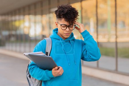 Foto de Introspectiva joven brasileño chico estudiante contempla mientras sostiene un cuaderno y usar una sudadera con capucha azul al aire libre - Imagen libre de derechos