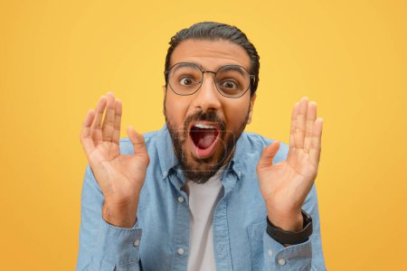 Overjoyed indian man with hands raised and an open mouth, exuding excitement on a yellow backdrop