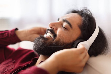 Photo for Relaxed Indian man in red enjoying music with white headphones while resting on a couch at home, closeup - Royalty Free Image