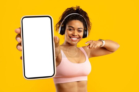 Photo for Smiling fit african american woman in a gym outfit shows a smartphone screen with a blank display, pointing to it, isolated on yellow - Royalty Free Image