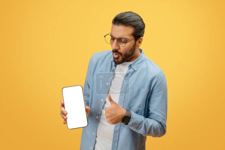 Foto de Un hombre indio sorprendido apunta a un teléfono inteligente con una pantalla en blanco, adecuada para la publicidad, sobre un fondo amarillo - Imagen libre de derechos