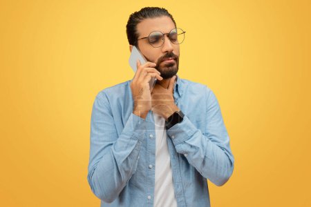 Téléchargez les photos : Un Indien barbu à lunettes et une chemise bleue discute avec un médecin sur son smartphone, touchant la gorge - en image libre de droit