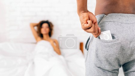 Représente un homme afro-américain tenant discrètement un préservatif dans sa poche avec une femme au lit ignorant en arrière-plan