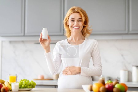 Foto de Alegre madre embarazada sosteniendo una botella de vitaminas prenatales en una cocina bien surtida - Imagen libre de derechos