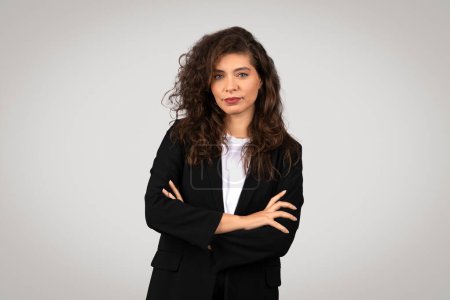 Eine professionelle Frau mit lockigem Haar in Businesskleidung mit selbstbewusster Haltung und neutralem Hintergrund