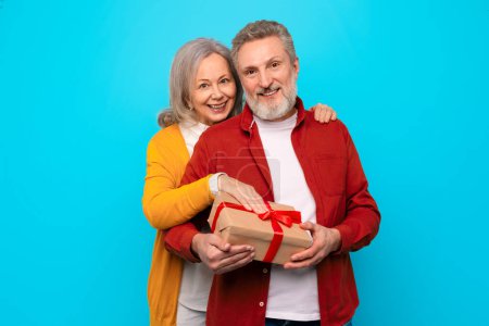 Téléchargez les photos : Aimer les conjoints mûrs européens embrassant tout en tenant boîte cadeau enveloppé sur fond bleu studio, souriant à la caméra profiter anniversaire de la relation ou célébration de la Saint-Valentin - en image libre de droit