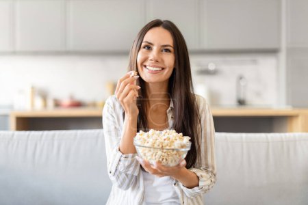 Casual brunette lady enjoying a bowl of popcorn in a contemporary kitchen, looking content with a smile