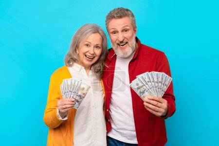Photo for Retirement prosperity. Smiling senior man and woman holding dollar money cash against blue studio backdrop, sharing excitement of victory and success. Financial safety and wealth - Royalty Free Image