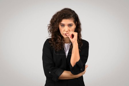 Photo for A woman with curly hair looks pensive, biting her nail, wearing a professional black suit against a grey backdrop - Royalty Free Image