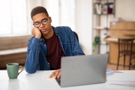 Adolescent ennuyé portant des lunettes montre l'ennui tout en utilisant un ordinateur portable à un bureau à domicile avec une tasse de café