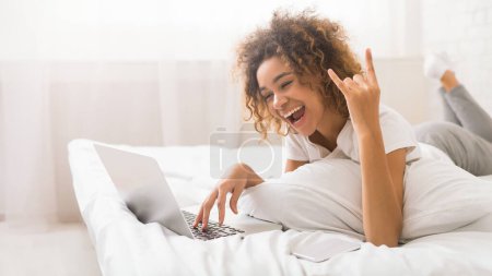 Una mujer afroamericana relajada trabaja en su computadora portátil desde la comodidad de su cama mientras parpadea una señal de paz con su mano