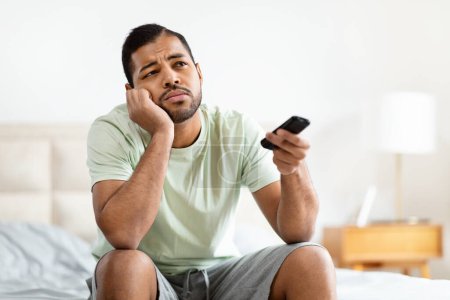 Téléchargez les photos : Un homme afro-américain peu impressionné tient une télécommande de télévision, indiquant l'ennui ou l'insatisfaction tout en étant assis sur son lit - en image libre de droit