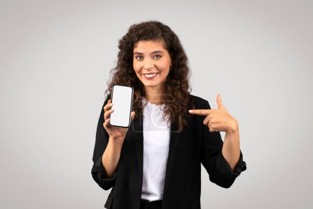 Foto de Una mujer alegre en traje de negocios sosteniendo un teléfono inteligente y apuntando a su pantalla en blanco, invitando a ver - Imagen libre de derechos