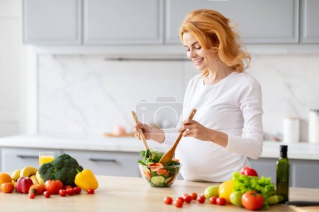 Eine blonde schwangere Frau mixt einen Salat, umgeben von frischem rohem Gemüse in einer modernen Küche