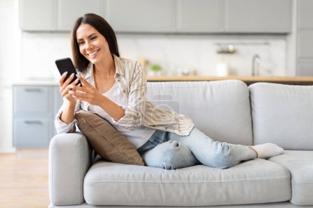 Eine Frau lümmelt auf einem Sofa und lächelt, während sie in einem gut beleuchteten, gemütlichen Raum zu Hause auf ihr Smartphone blickt.
