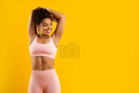 Foto de Una alegre mujer afroamericana en ropa deportiva rosa posa con las manos en la cabeza sobre un vibrante telón de fondo amarillo, lo que indica salud y actividad - Imagen libre de derechos