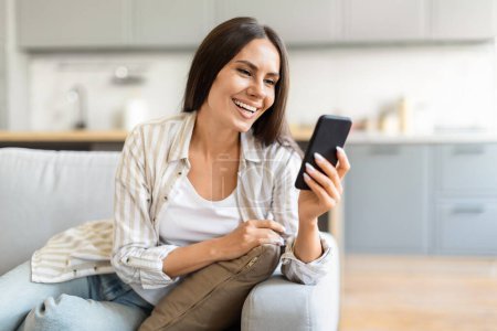Photo for Cheerfully seated on the couch, this woman texts on her phone, exhibiting an ease with digital communication - Royalty Free Image