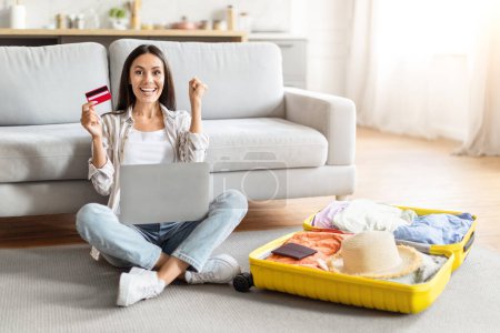 Mujer extática por hacer una compra en línea, con una tarjeta de crédito y un ordenador portátil, sentada entre artículos de viaje desempaquetados