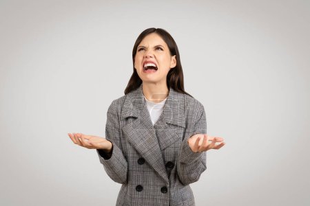 Photo for Angry businesswoman in gray houndstooth blazer, shouting with hands raised in exasperation, showing frustration or disagreement, on neutral background - Royalty Free Image