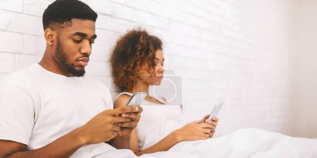 Afroamerikanisches Paar im Bett mit umgedrehtem Rücken und in ihre Smartphones vertieft, was moderne Beziehungen widerspiegelt