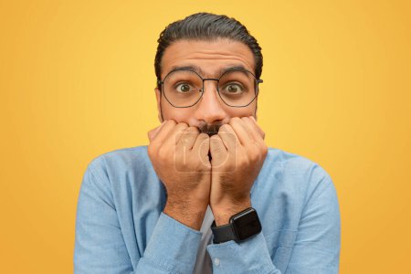 Biting nails, a worried indian man stands looking anxious against a yellow background, signifying tension