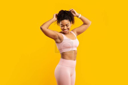 Téléchargez les photos : Une jeune femme afro-américaine dynamique en tenue d'entraînement rose prépare joyeusement ses cheveux pour une séance de fitness contre un mur jaune - en image libre de droit