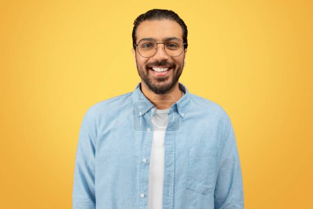 Un hombre indio alegre con gafas, una camisa azul y una camiseta blanca sonriendo sobre un soleado fondo amarillo