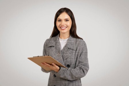 Téléchargez les photos : Jeune femme d'affaires confiante tenant le presse-papiers, prête à se rencontrer, portant un blazer intelligent à dents de poule et souriant à la caméra, sur fond gris - en image libre de droit