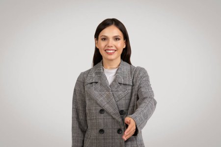 Photo for Cheerful young businesswoman extending friendly handshake, dressed in fashionable plaid blazer, against neutral gray background - Royalty Free Image