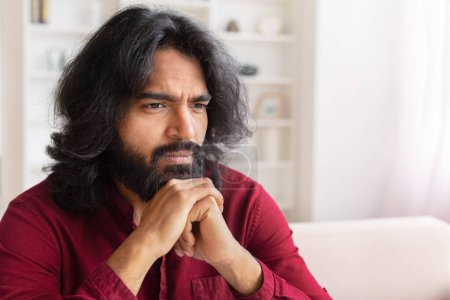Foto de Un hombre indio reflexivo apoya su barbilla en sus manos, contemplando en una habitación con un ambiente luminoso y luminoso - Imagen libre de derechos