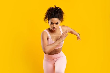 Téléchargez les photos : Une femme sportive afro-américaine concentrée en tenue de sport rose étire les bras, se préparant à une séance d'entraînement sur un fond jaune - en image libre de droit