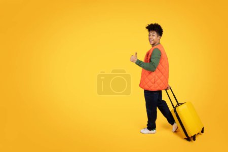 Foto de Un joven afroamericano alegre da un pulgar hacia arriba mientras camina con una maleta amarilla sobre un vibrante fondo amarillo - Imagen libre de derechos